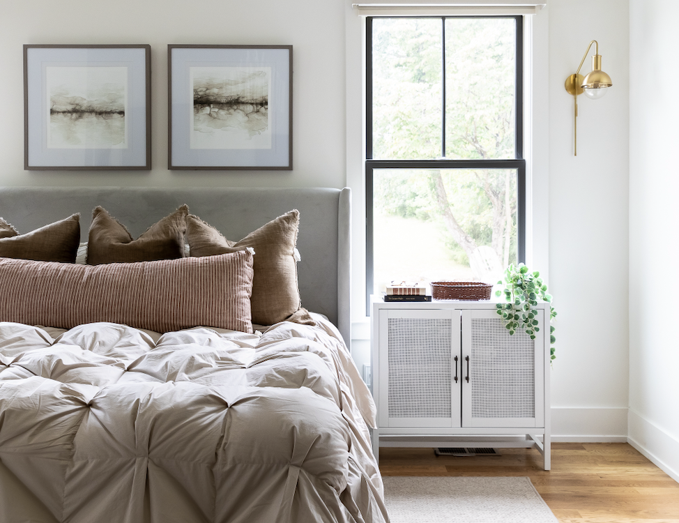 A neatly made bed with brown pillows and a taupe comforter is positioned next to a window with a white cabinet underneath, featuring a hanging plant and wall-mounted lamp above.